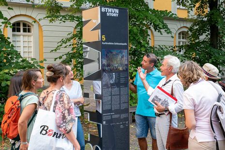 Eine Personengruppe steht an einer Stele des BTHVN-Rundgangs zu Ehren Ludwig van Beethovens