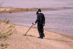 Zwei Männer suchen ein Flussufer mit einem Metalldetektor ab.