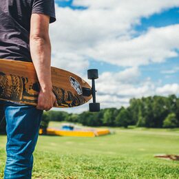 Ein junger Mann steht in der Rheinaue und hält ein Longboard unterm Arm.