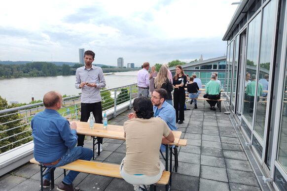 Gruppe von Personen im Außenbereich eines Restaurants am Rhein mit dem Siebengebierge im Hintergrunde. Die Personen diskutieren.