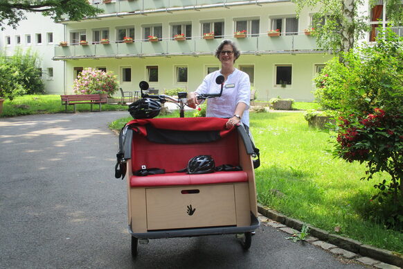 Eine Frau steht im Garten vor einer Hausfassade und zeigt eine Rikscha.