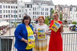 Ina Harder, Katja Dörner und Patty Burgunder halten auf dem Beueler Rathausbalkon das bunte Plakat des Abschlusskonzertes zum Jubiläumsjahr "200 Jahre Beueler Weiberfastnacht" in Händen. Im Hintergrund der Möhneplatz und die Friedrich-Breuer-Straße.