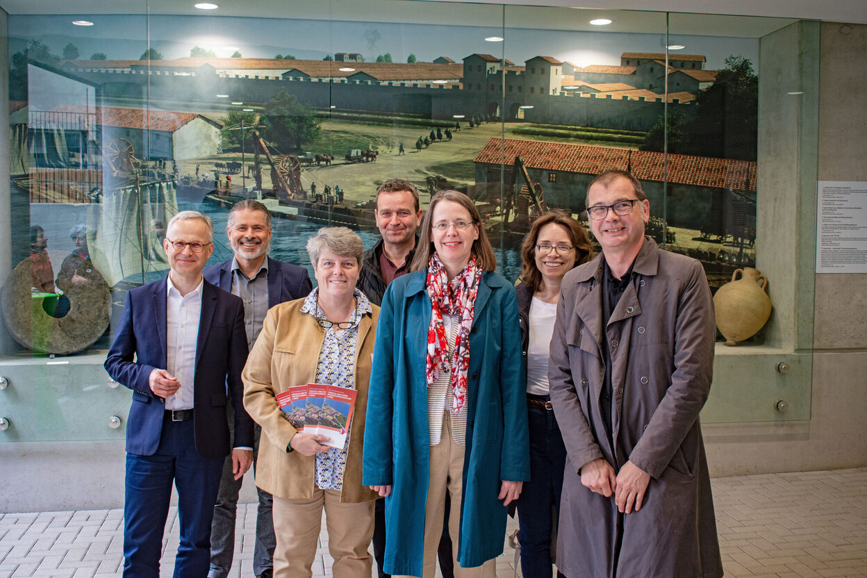 Gruppenbild vor einem großformatigen Schaukasten mit einer Zeichnung des ehemaligen Römerlagers in Bonn.