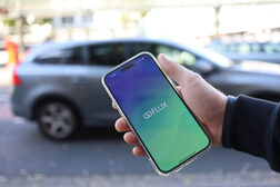 Eine Hand hält ein Smartphone mit der GoFlux-App, im Hintergrund ist unscharf ein Auto zu erkennen