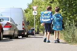 Zwei Jungen mit großen Schulranzen auf einem Gehweg neben geparkten Autos