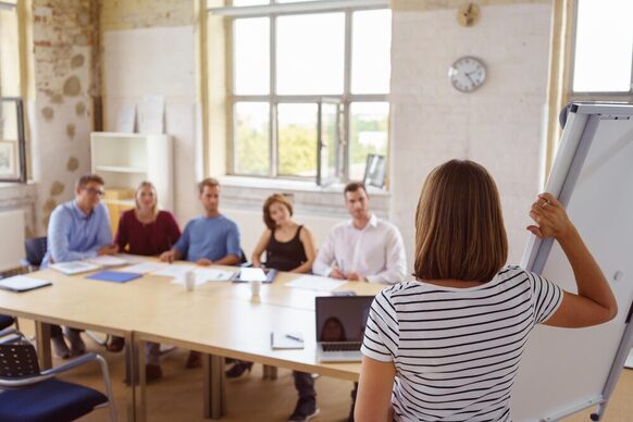 Eine Frau steht an einem Flipchart und hält vor einer Gruppe von Kolleginnen und Kollegen eine Präsentation