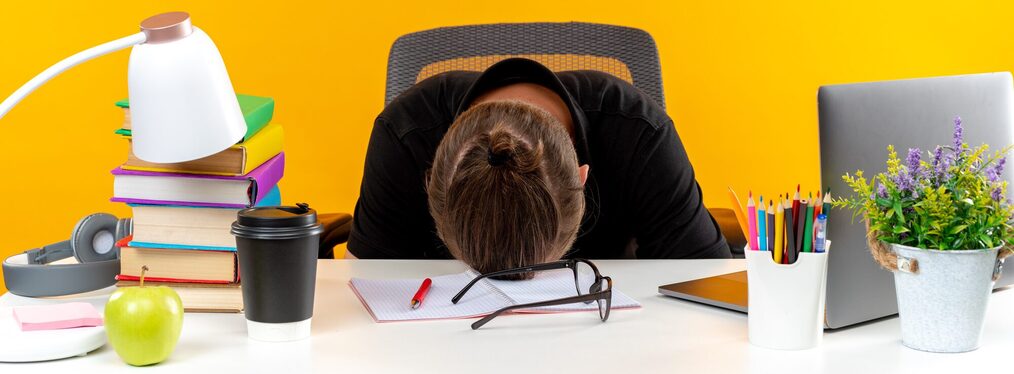 young guy student sitting at table with school tools lowered head on table isolated on orange background