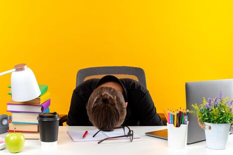 young guy student sitting at table with school tools lowered head on table isolated on orange background