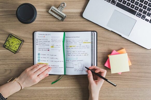 Hände halten ein aufgeschlagenes Notizbuch und einen Stift, im Hintergrund steht ein Laptop