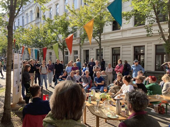 Festlich geschmückte Straße mit Bürger*innen, die im Freien einem Vortrag lauschen.