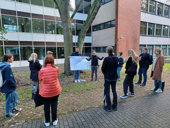 Eine Gruppe Menschen stehen im Halbkreis vor zwei Personen, die mithilfe eines Plakats etwas erklären.